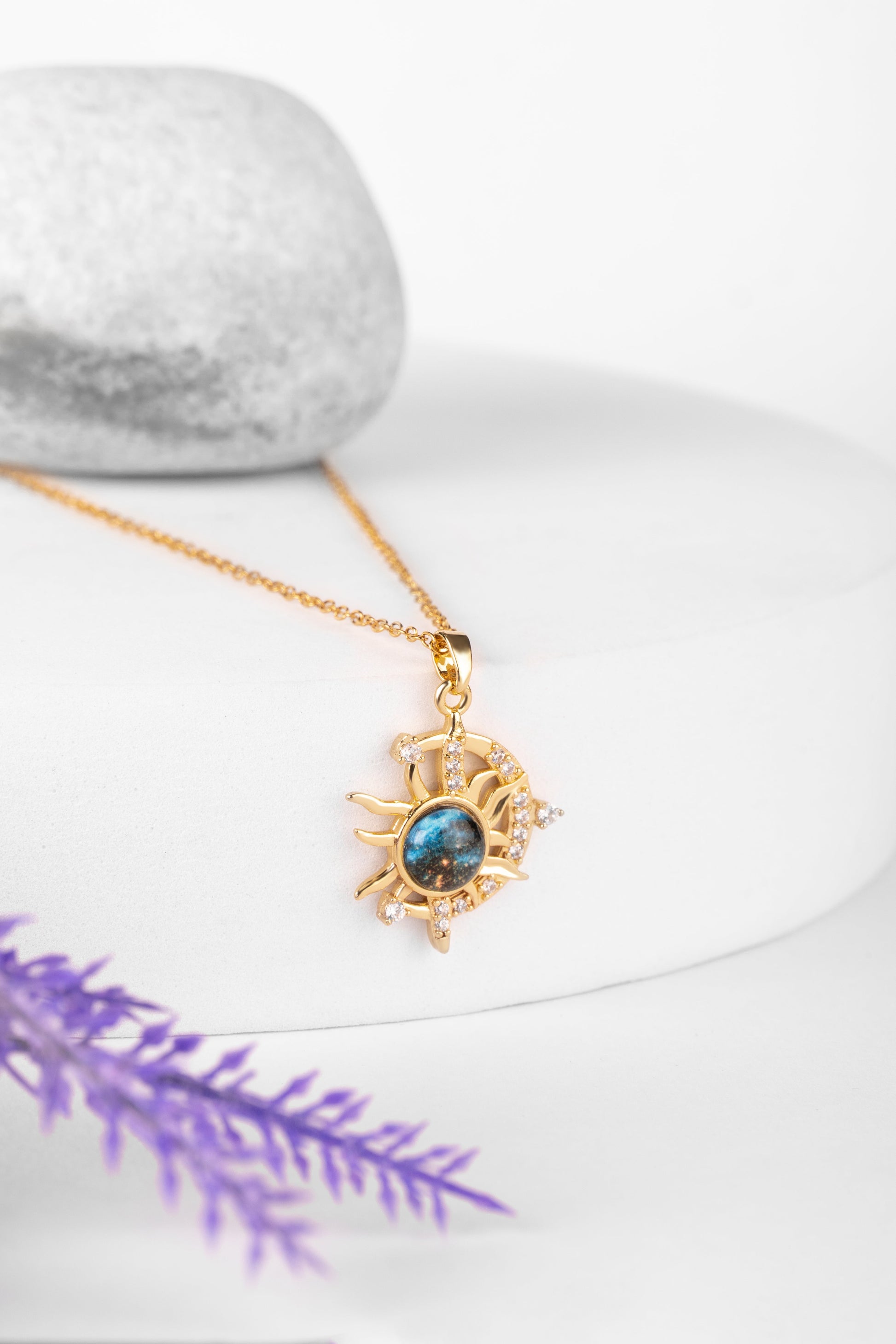 Gold necklace with a sun-shaped pendant on a white surface with purple leaves.
