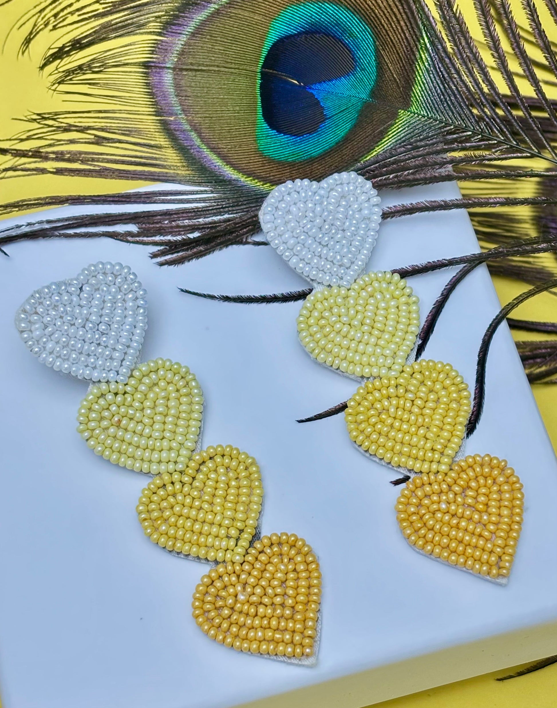 Heart-shaped earrings with yellow, green, and white beads on a peacock feather background