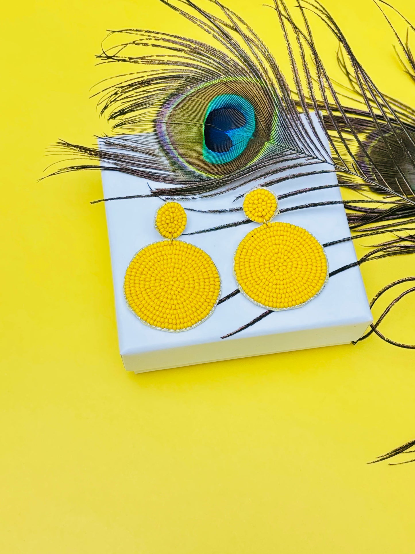 Yellow earrings on a white box with a peacock feather on a yellow background