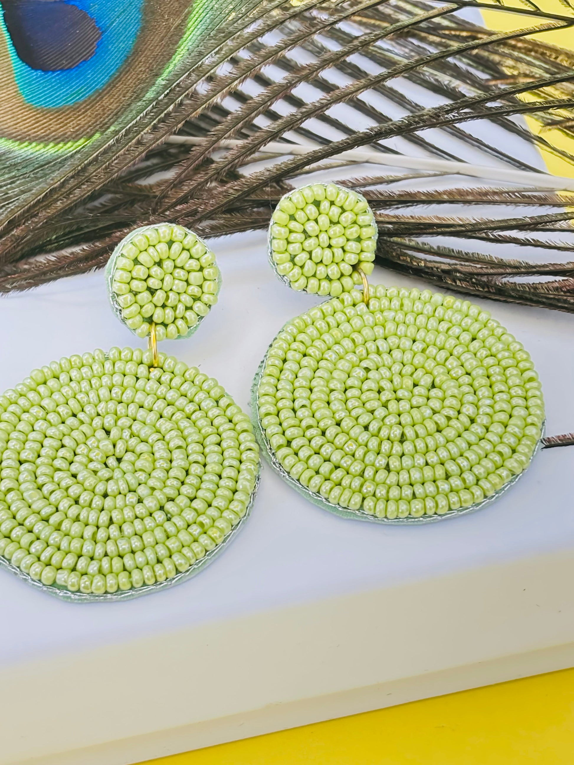Green beaded earrings on a white surface with peacock feathers in the background
