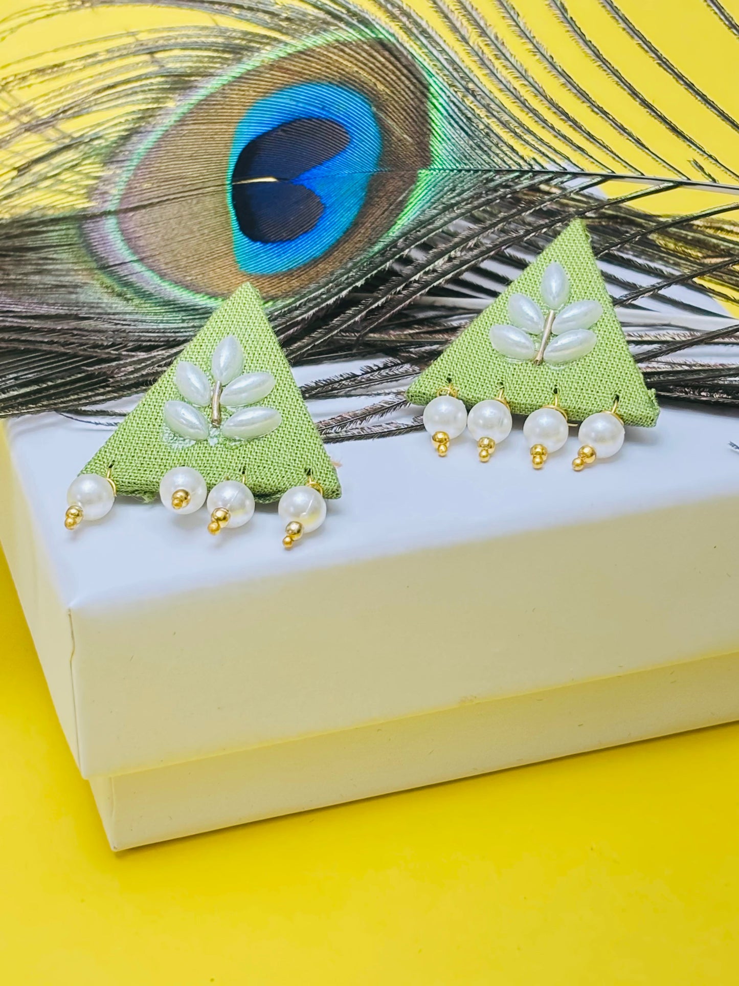 Green triangle-shaped earrings with pearl details on a peacock feather background