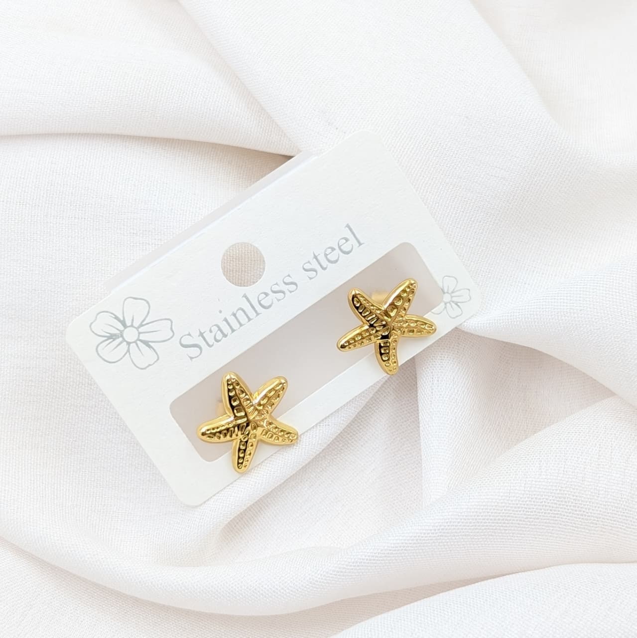Gold starfish earrings on a white card with 'Stainless steel' text, against a white fabric background.
