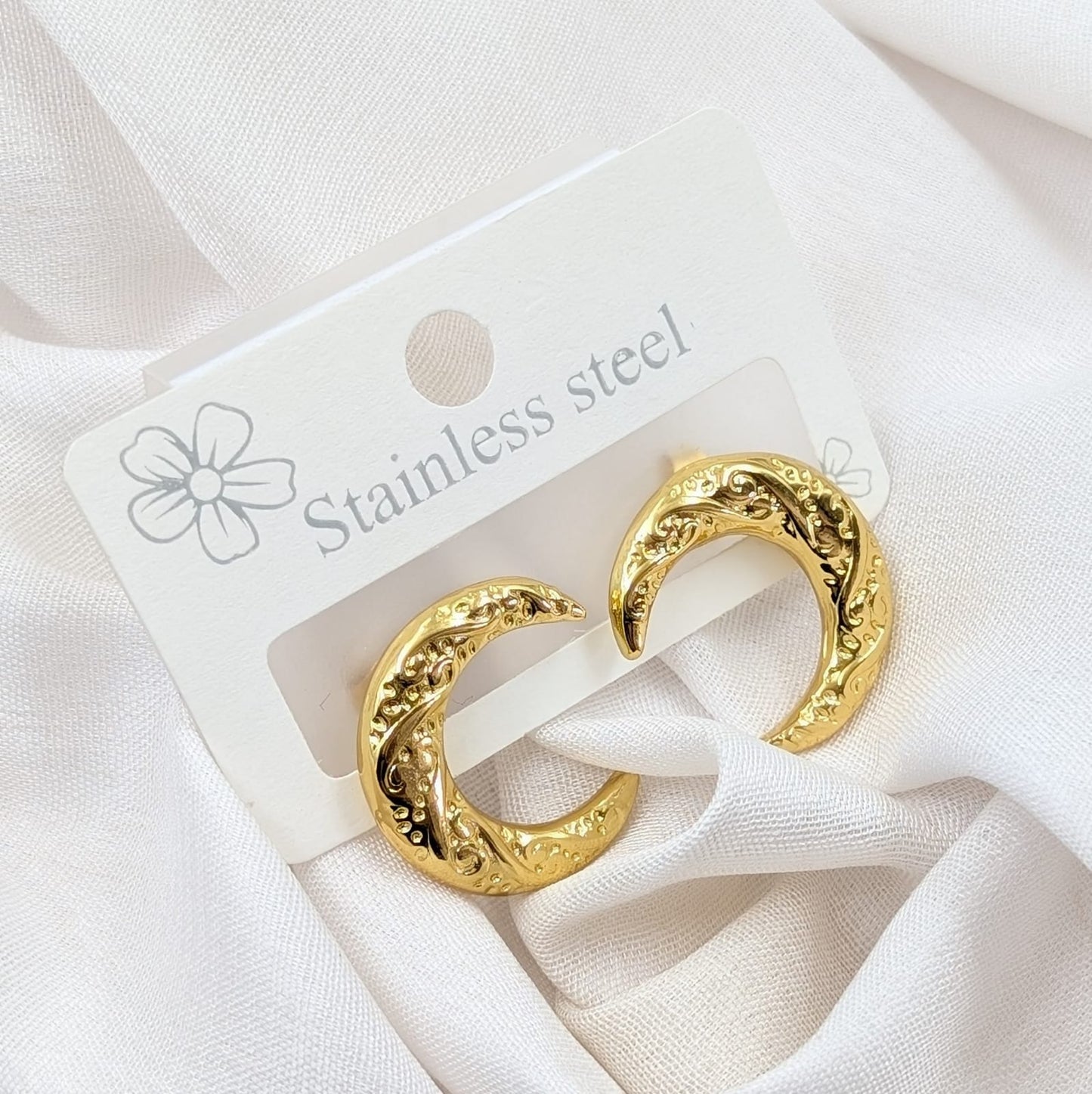 Gold hoop earrings on a white card with 'Stainless steel' text, placed on a white fabric background.