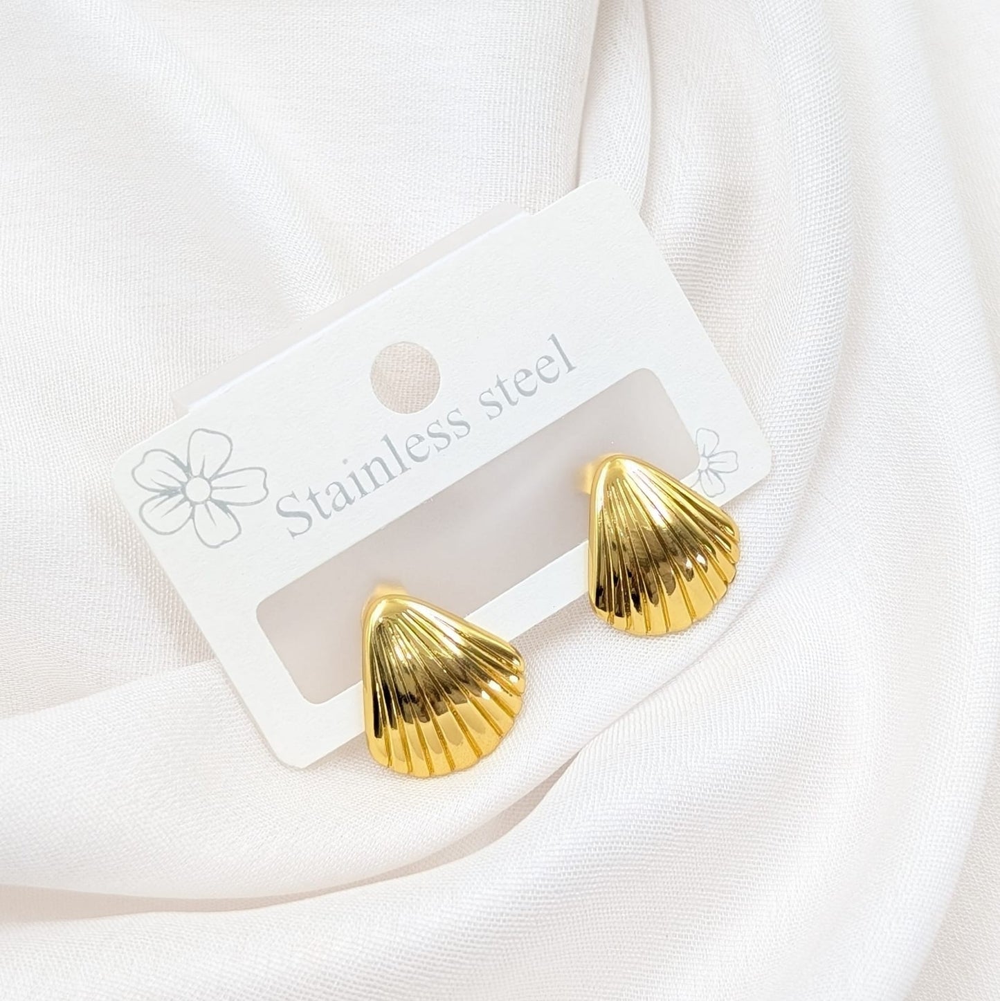 Gold shell-shaped earrings on a white card with 'Stainless steel' text, against a white fabric background.