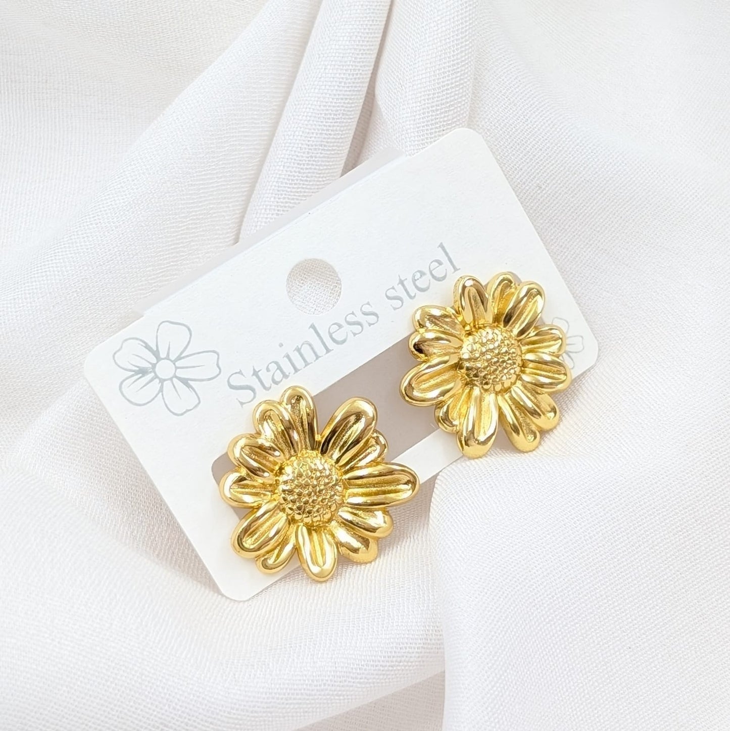 Gold flower-shaped earrings on a white card with 'Stainless steel' text, against a white fabric background.