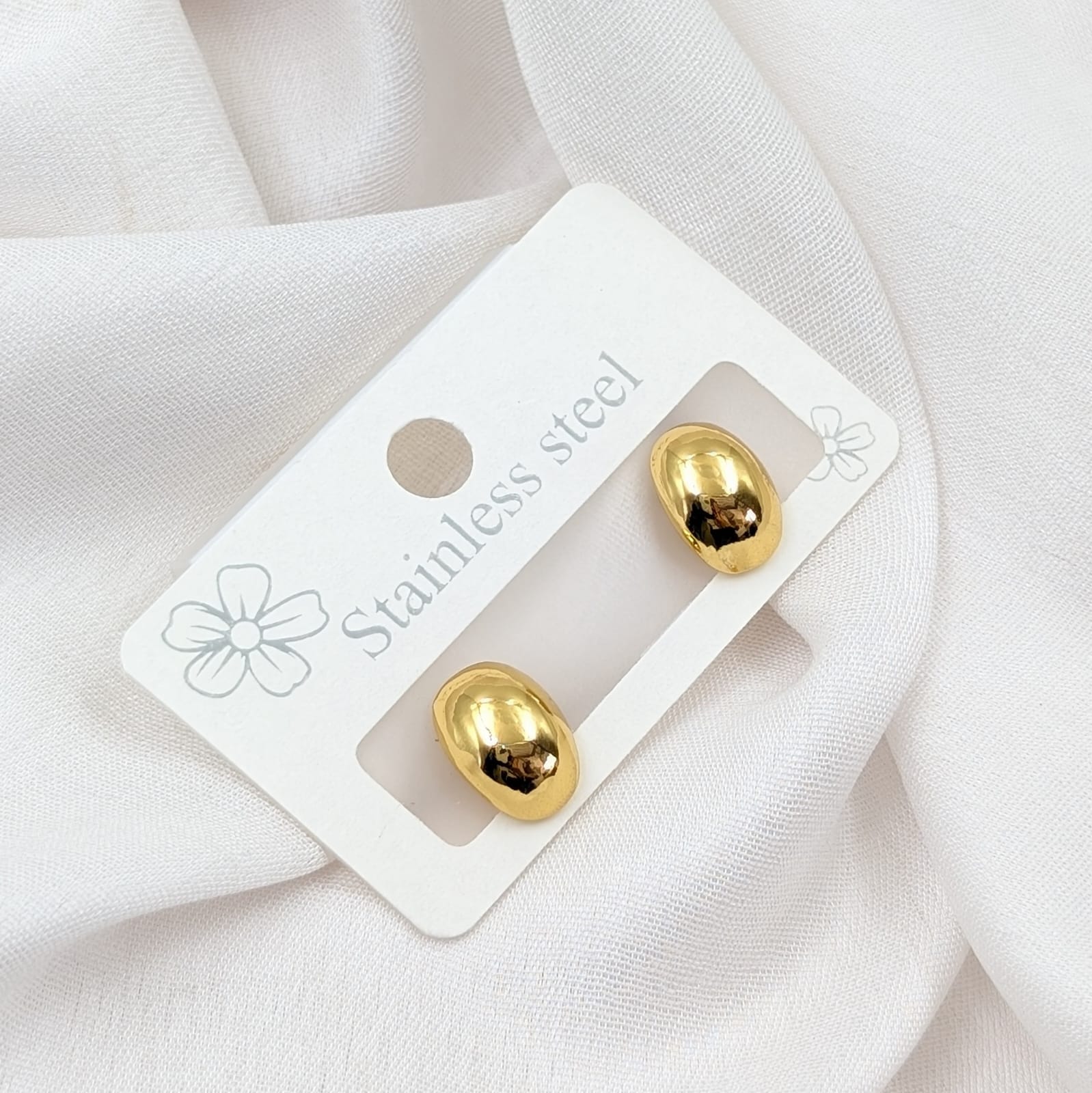 Gold earrings on a white card with 'Stainless steel' text, placed on a white fabric background.