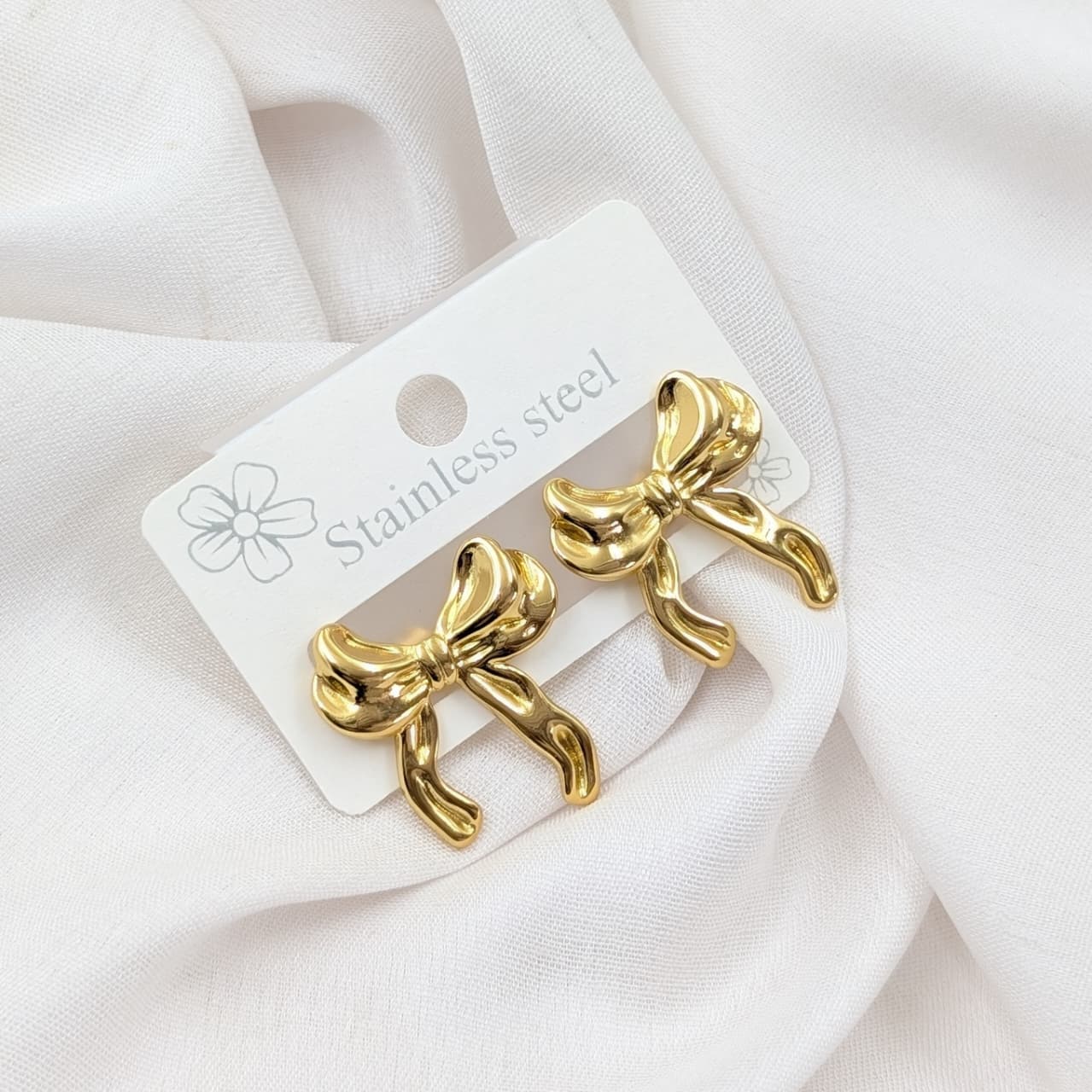 Gold bow-shaped earrings on a white card with 'Stainless steel' text, placed on a white fabric background.